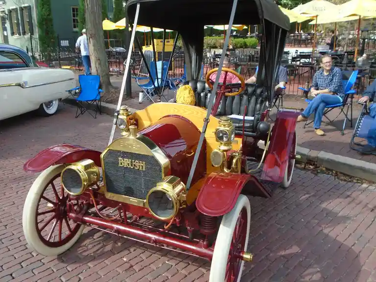A vintage red and brass Brush automobile with wooden accents and white-walled tires is parked on a brick street, surrounded…