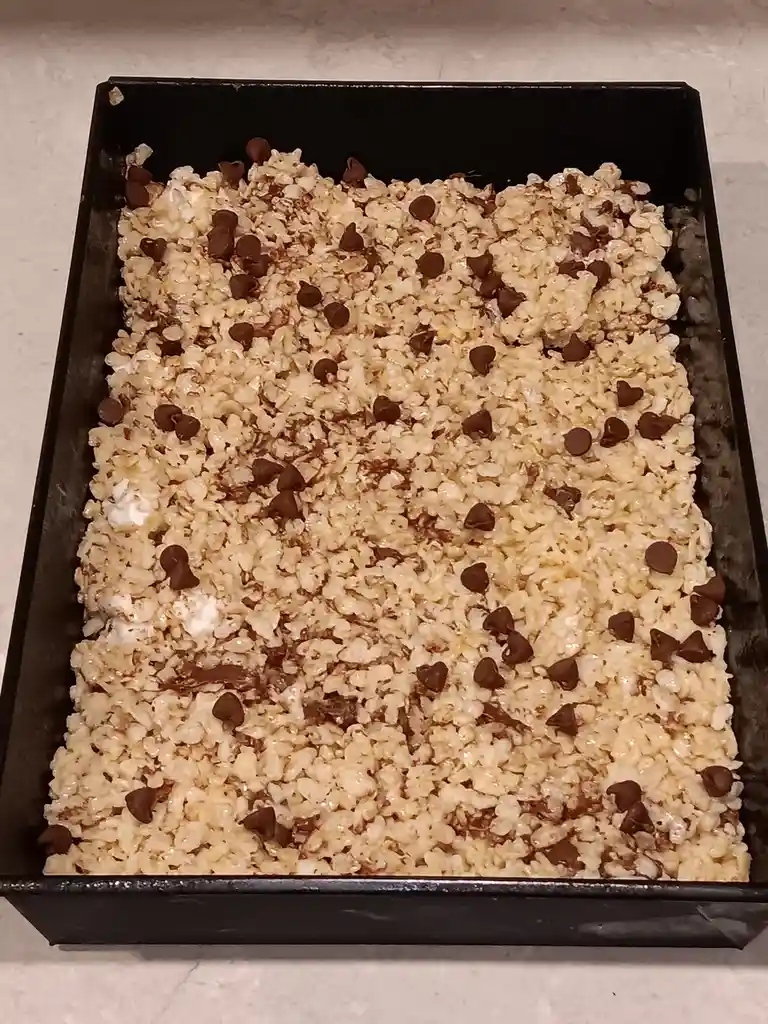 A rectangular black baking pan contains uncooked rice krispie treats mixed with chocolate chips, arranged to resemble a sad…