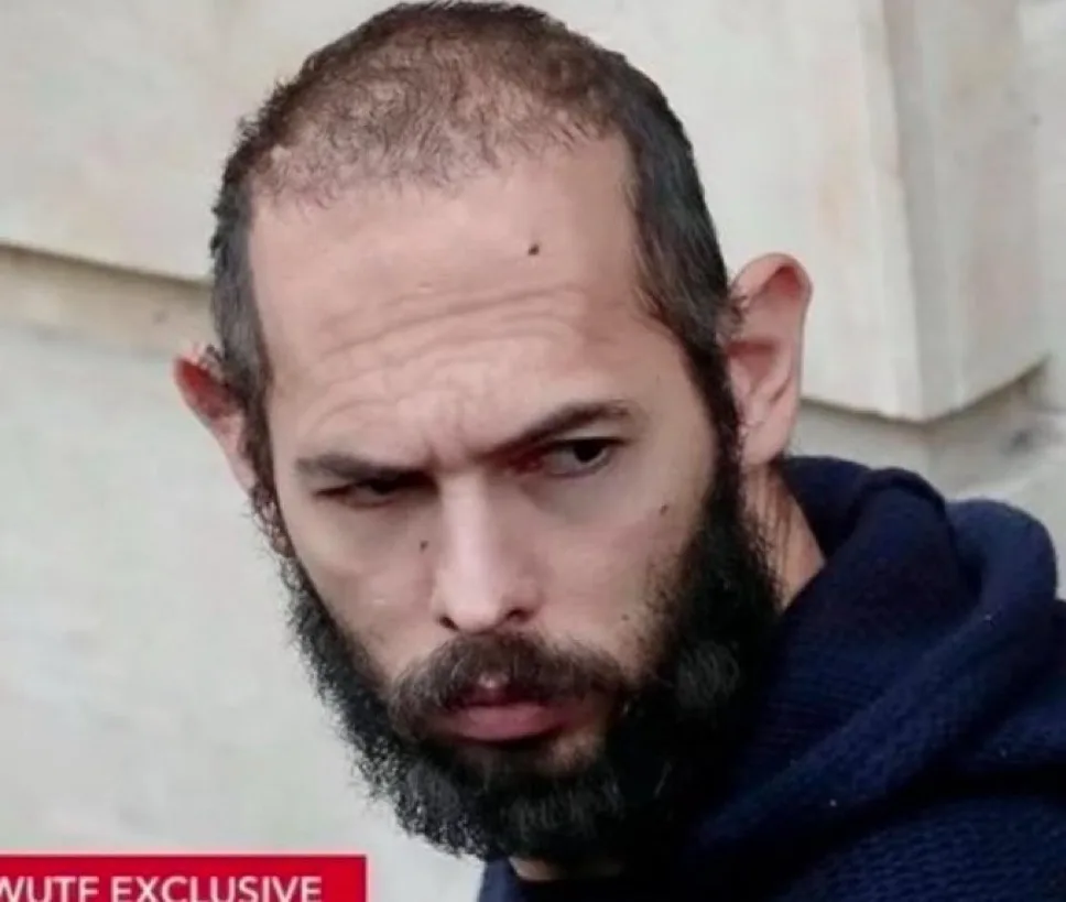 A close-up photo of a man with a receding hairline, a full beard, and a skeptical, annoyed expression, wearing a dark blue…