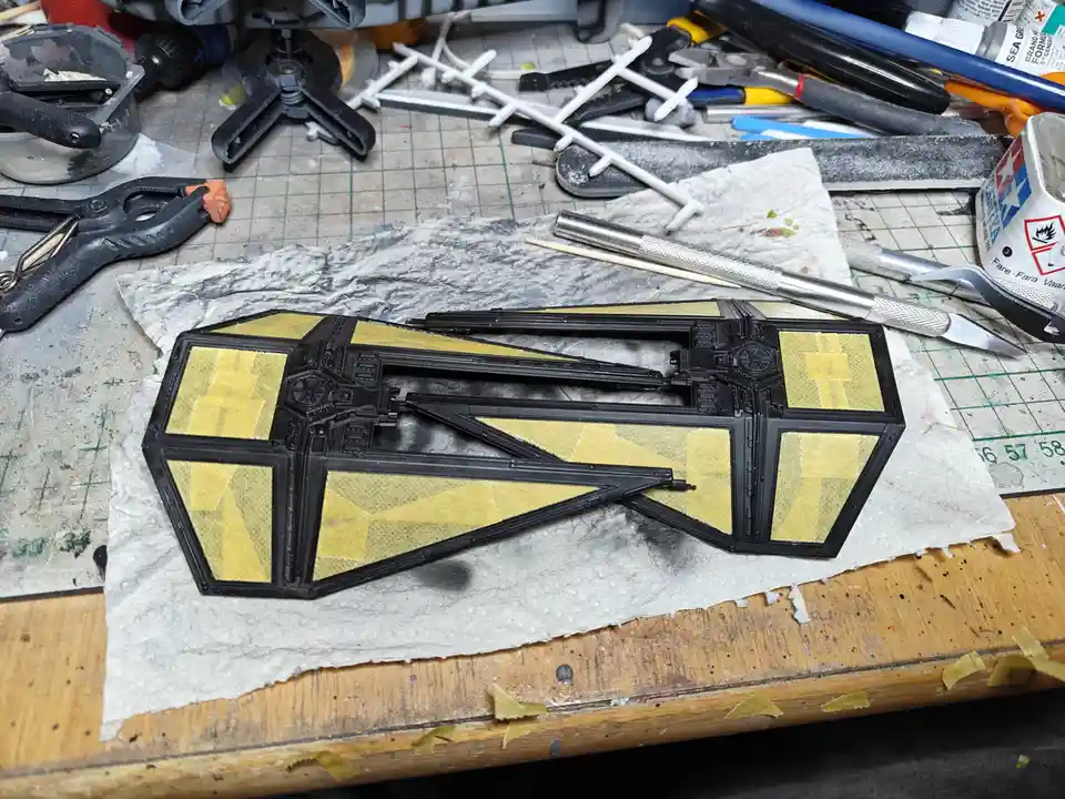 A partially painted black plastic Star Destroyer model sits on a workbench, with yellow paint applied in rough, uneven…