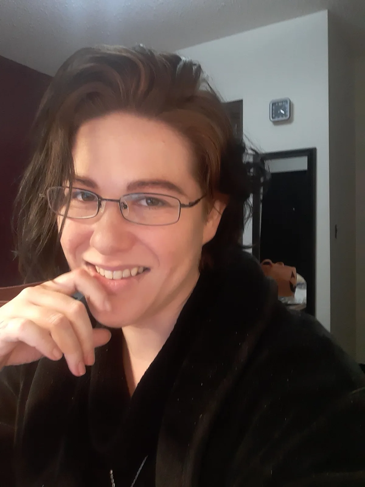 A person with glasses and dark hair, wearing a black robe, smiles while resting their chin on their hand. In the background…