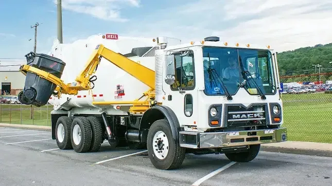 A white Mack garbage truck with a yellow mechanical arm is shown lifting a black trash can. The word 'HELL' is visible in…