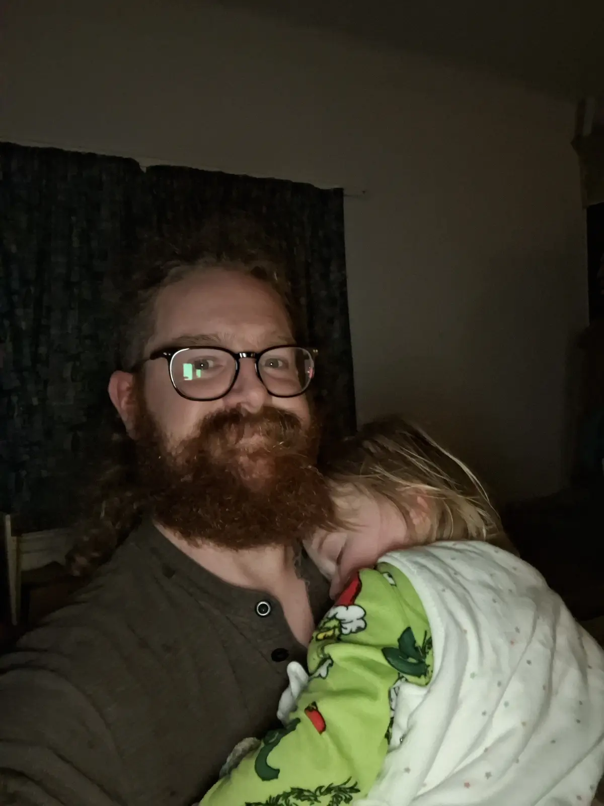A man with a full beard and glasses holds a sleeping child in a green and white pajama outfit. The man looks directly at the…