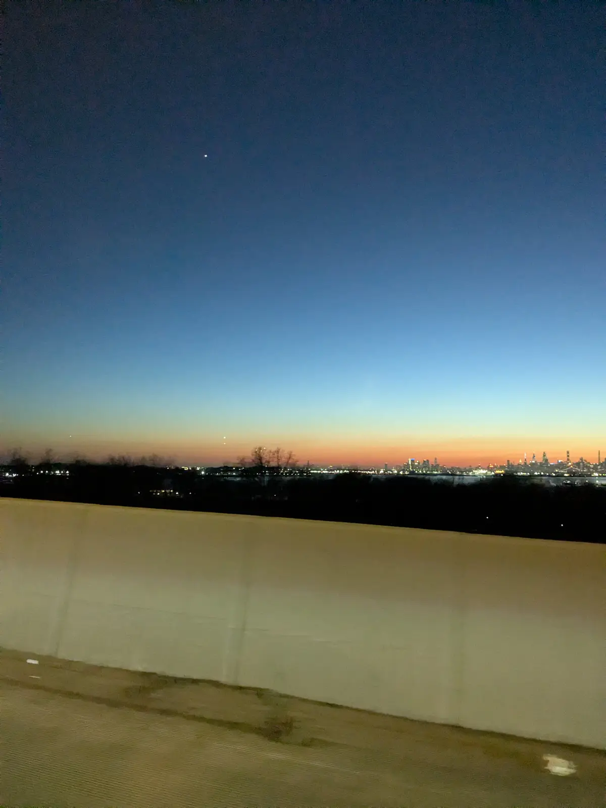 A photo taken from a moving vehicle at dusk shows a city skyline glowing on the horizon under a gradient sky transitioning…