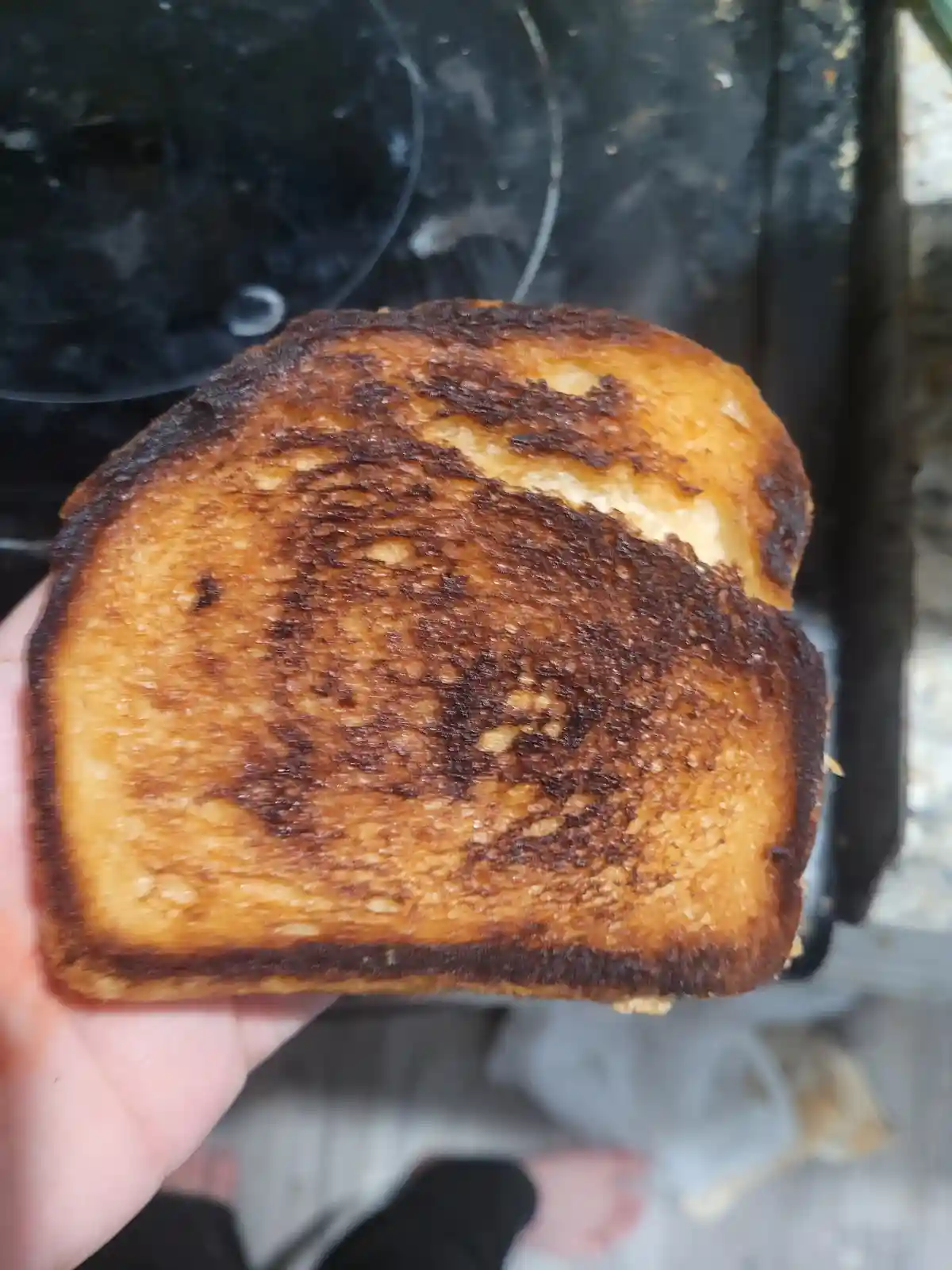 A hand holds up a slice of toast that is severely burnt, with dark char marks covering most of its surface, while the…