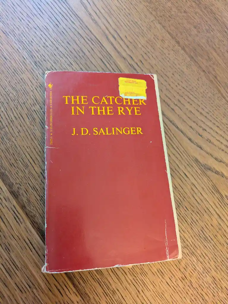 A well-worn red paperback copy of J.D. Salinger's 'The Catcher in the Rye' rests on a wooden surface, with a yellow 'USED'…