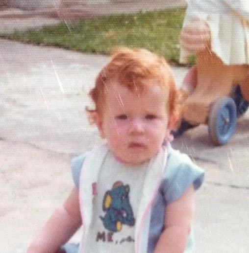 A grainy, vintage photo of a young child with reddish hair and a bib featuring a cartoon elephant, looking directly at the…