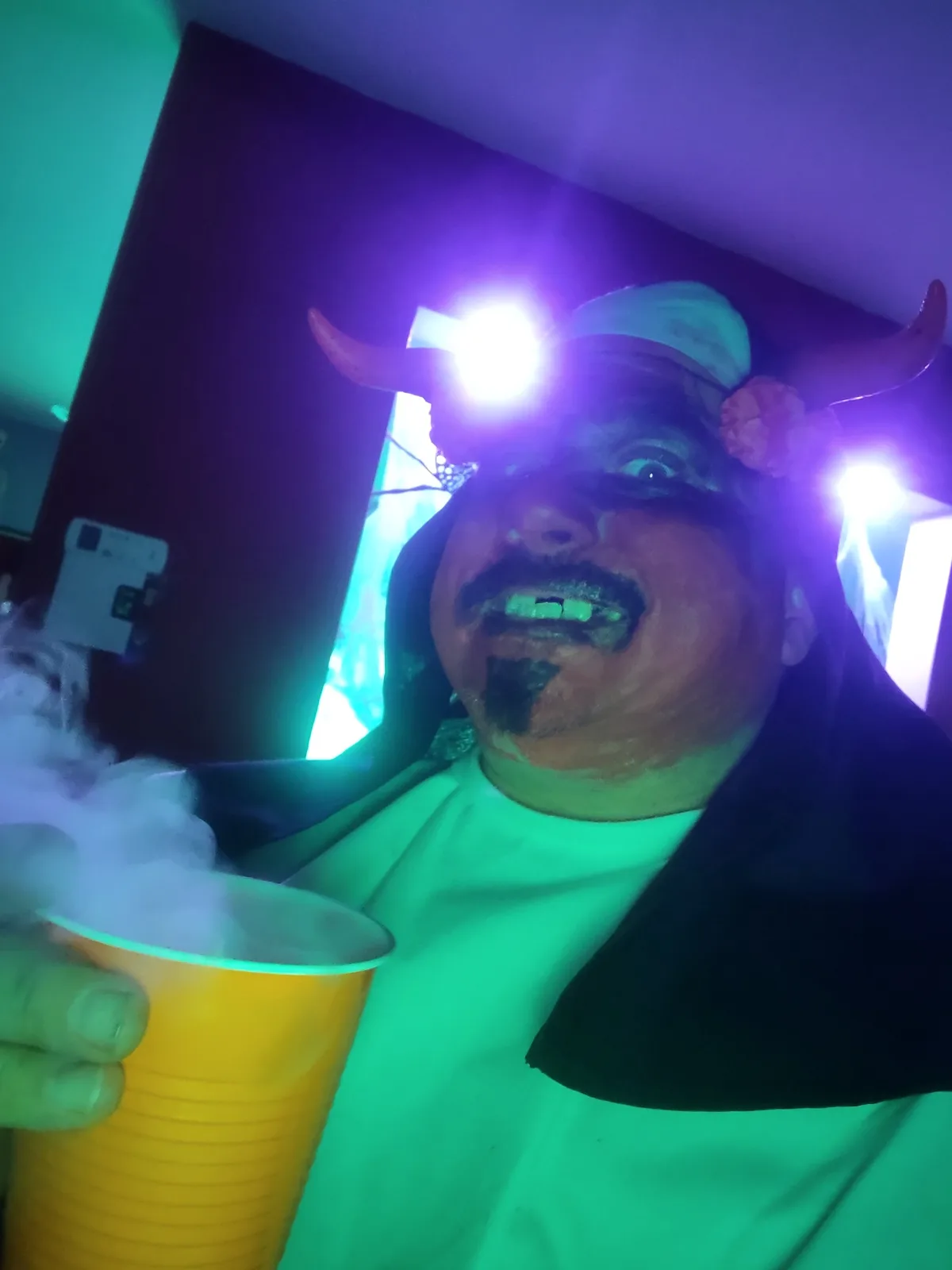 A man in a devil costume with horns and dark makeup, holding a yellow cup emitting smoke, under colorful party lights. The…