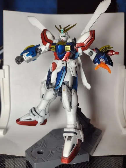 A detailed plastic model of a Gundam robot, primarily white with red and blue accents, stands in a dynamic pose on a grey…