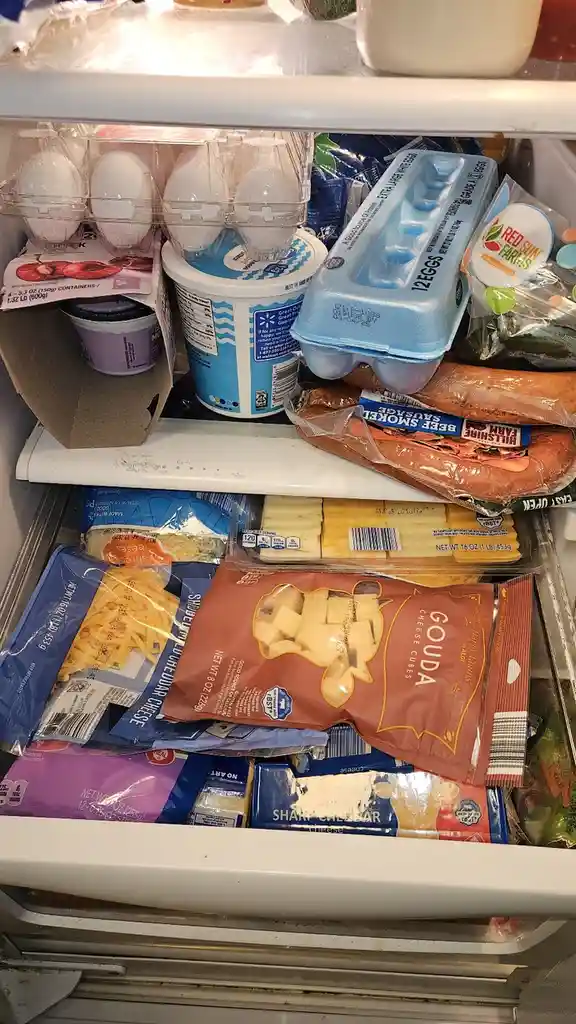 A close-up photo of a refrigerator's cheese drawer and upper shelf, overflowing with various cheese products including gouda…