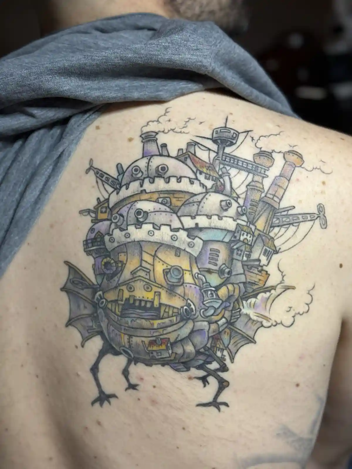 A close-up photo shows a detailed tattoo on a person's upper back, depicting the iconic flying castle from the animated film…