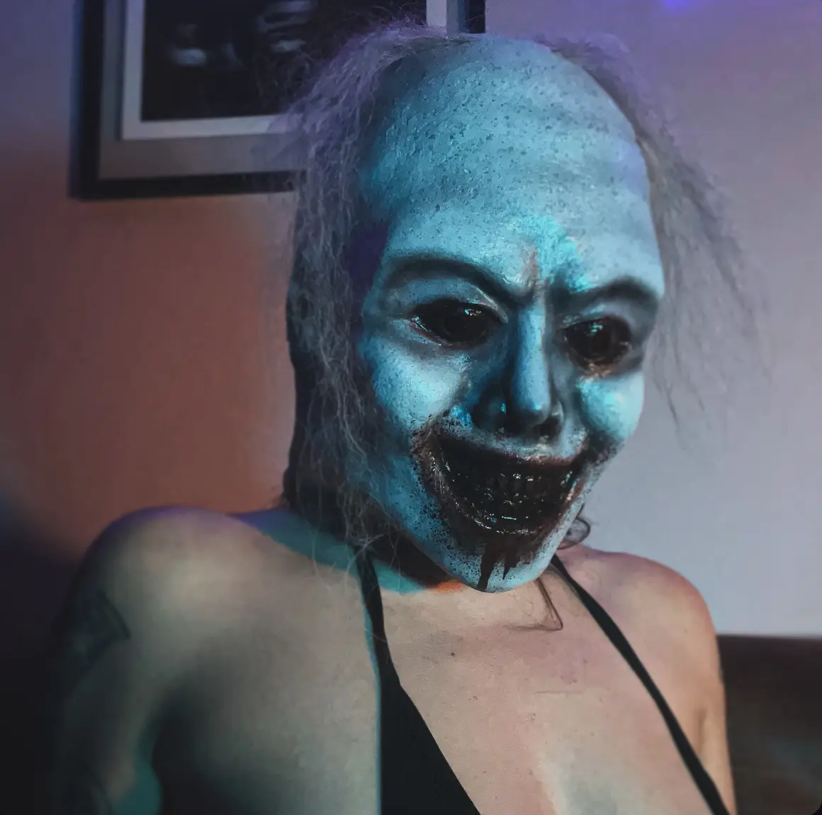 A person with pale blue face paint, darkened eyes, and fake blood dripping from a wide, toothy grin, wearing a black top…