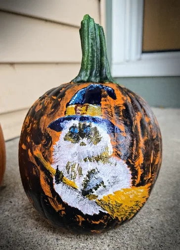 A hand-painted pumpkin with a witch face, featuring a blue hat, white face, and yellow broomstick, sits on a porch step. The…