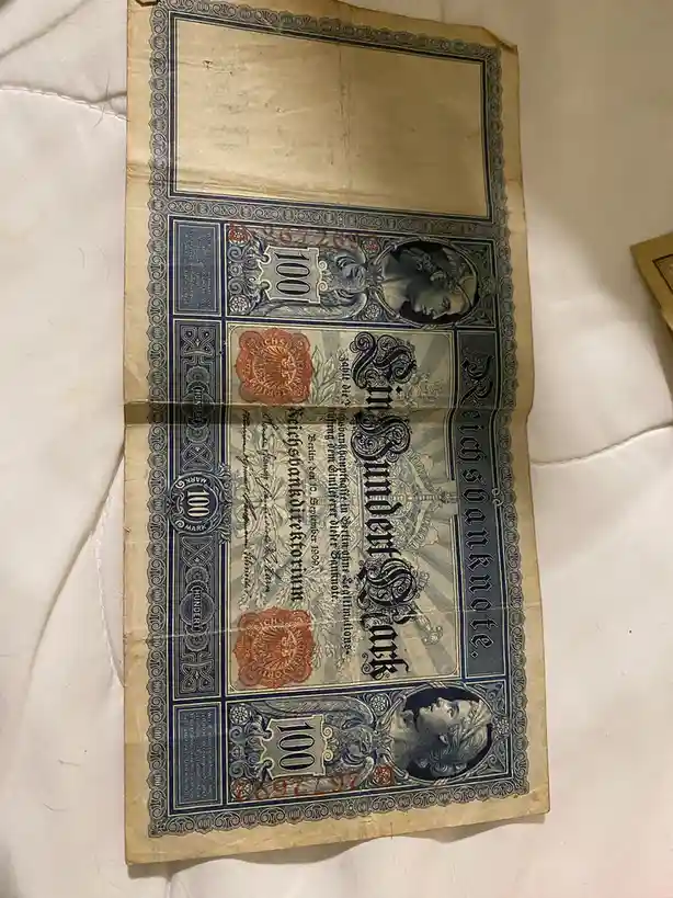 A folded, aged 100-mark German Reichsbanknote from 1909 lies on a white surface, its ornate blue and red design showing…