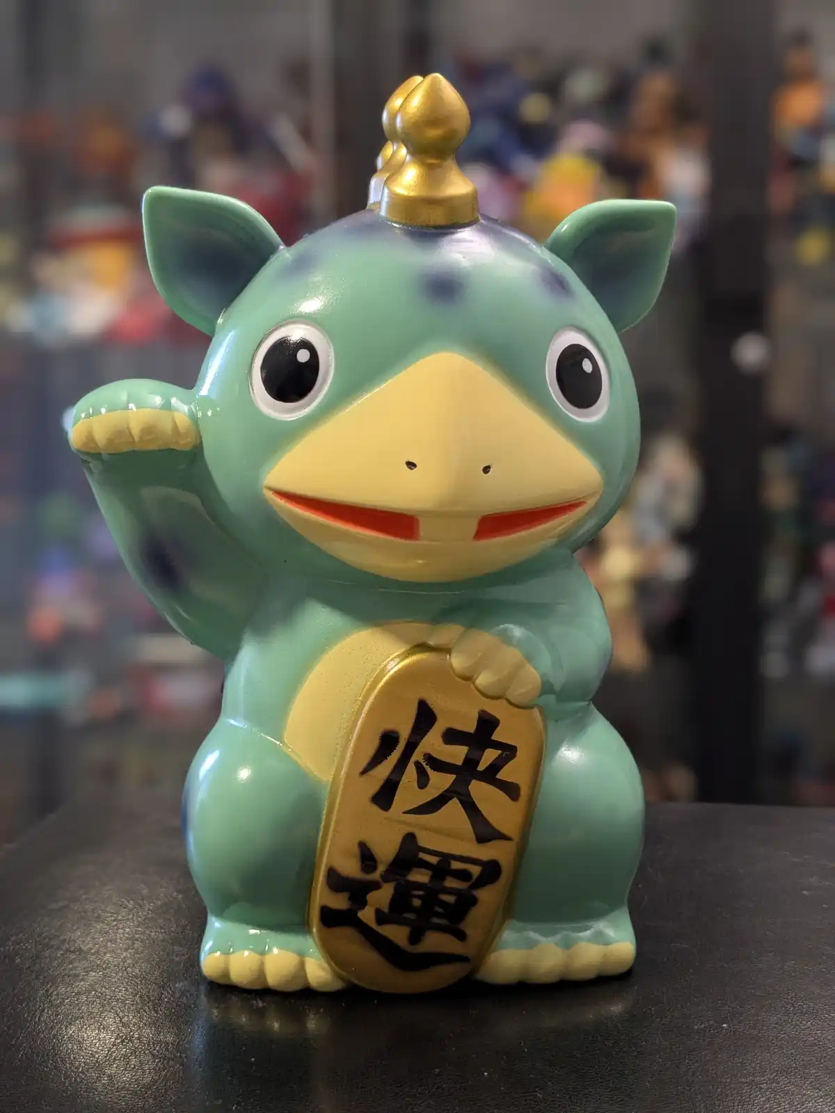 A green, cartoonish frog-like figurine with a gold crown and yellow beak, waving one hand while holding a golden plaque with…