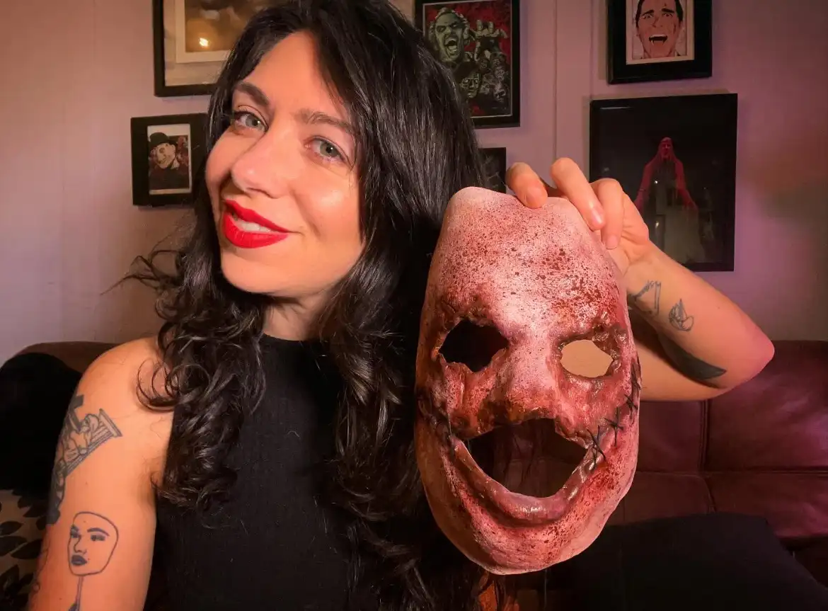 A woman with dark hair and red lipstick smiles while holding up a gruesome, blood-splattered horror mask with stitched eyes…
