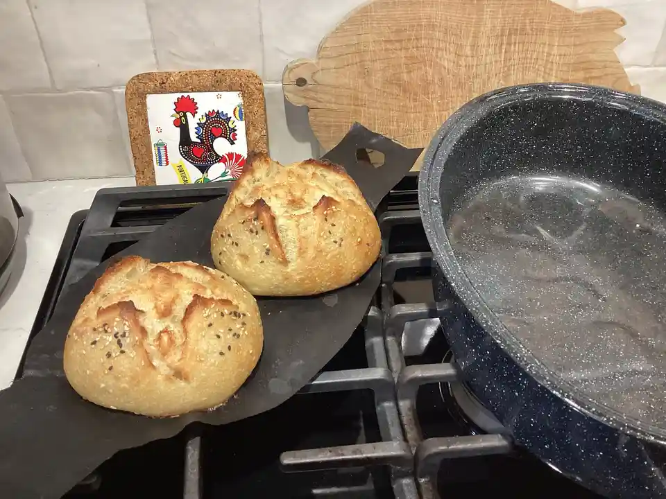 Two golden-brown, sesame-seeded artisan bread rolls sit on a black baking sheet placed directly on a gas stove burner. To…