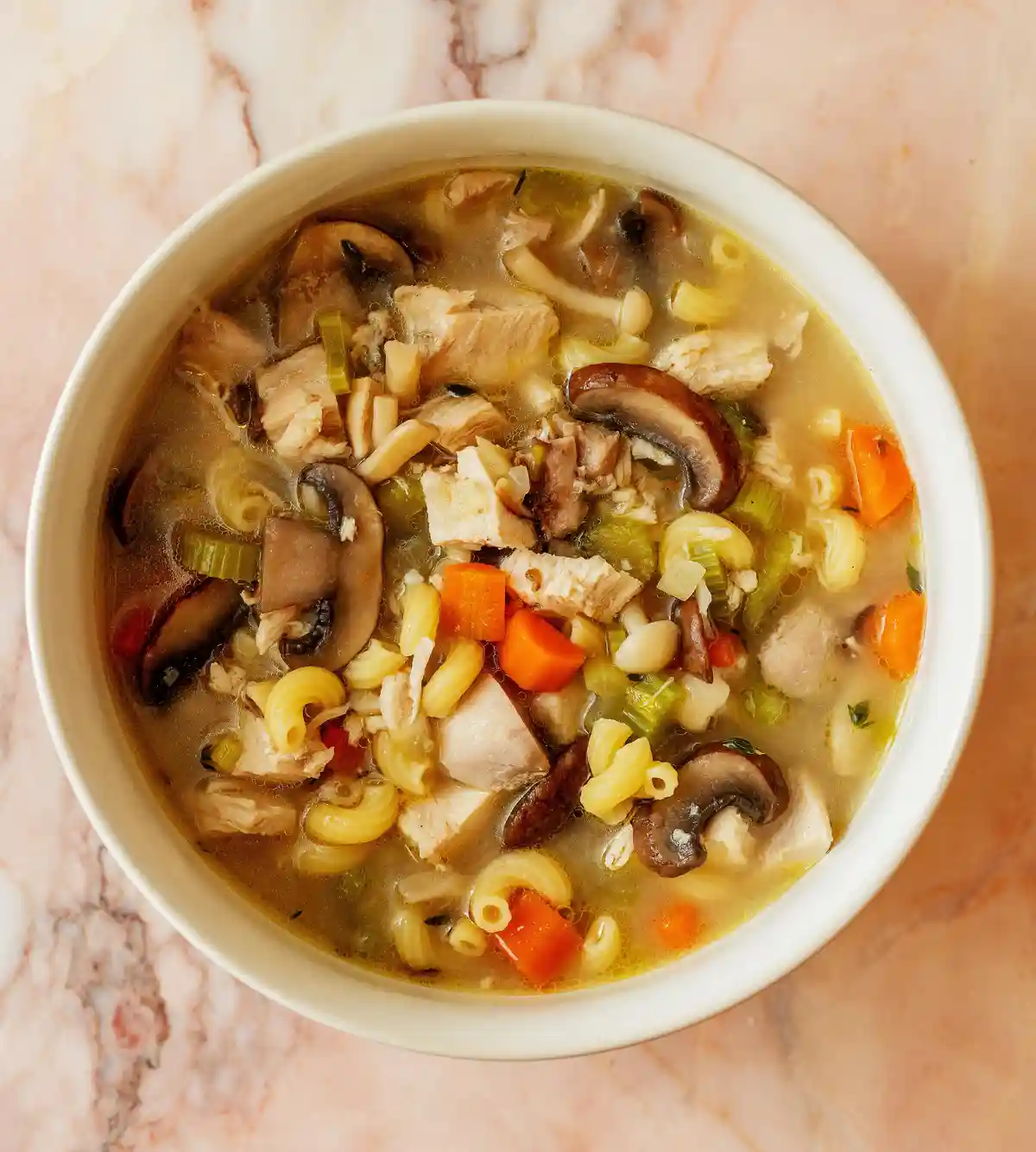 A top-down photo shows a white bowl filled with chicken noodle soup, containing visible chunks of chicken, sliced mushrooms…