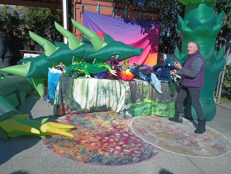 A woman stands beside a large green inflatable dragon at an outdoor market stall, holding a phone. The dragon's head and…