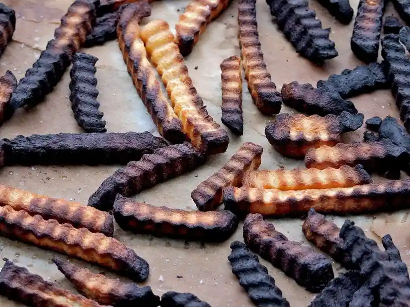 A close-up photo shows crinkle-cut french fries on parchment paper, with most of them burnt black and only a few slightly…