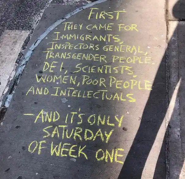 Yellow chalk writing on a dark concrete sidewalk spells out a satirical adaptation of the 'they came for...' quote, listing…