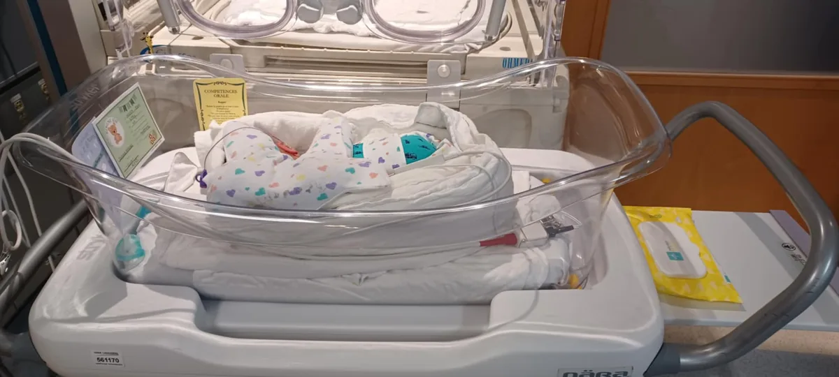 A newborn baby is swaddled in a white blanket with colorful hearts and lies in a clear plastic hospital bassinet. Medical…
