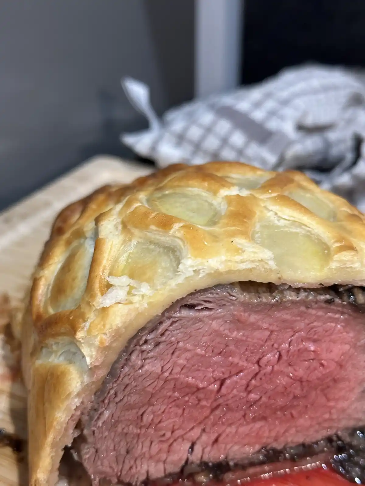 A close-up photo shows a perfectly sliced beef Wellington with a golden-brown, flaky puff pastry crust and a juicy…
