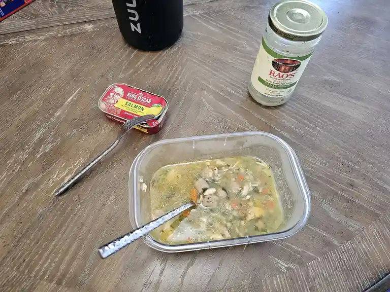A clear plastic container with mostly eaten soup sits on a wooden table, next to an open can of King Oscar salmon, a jar of…