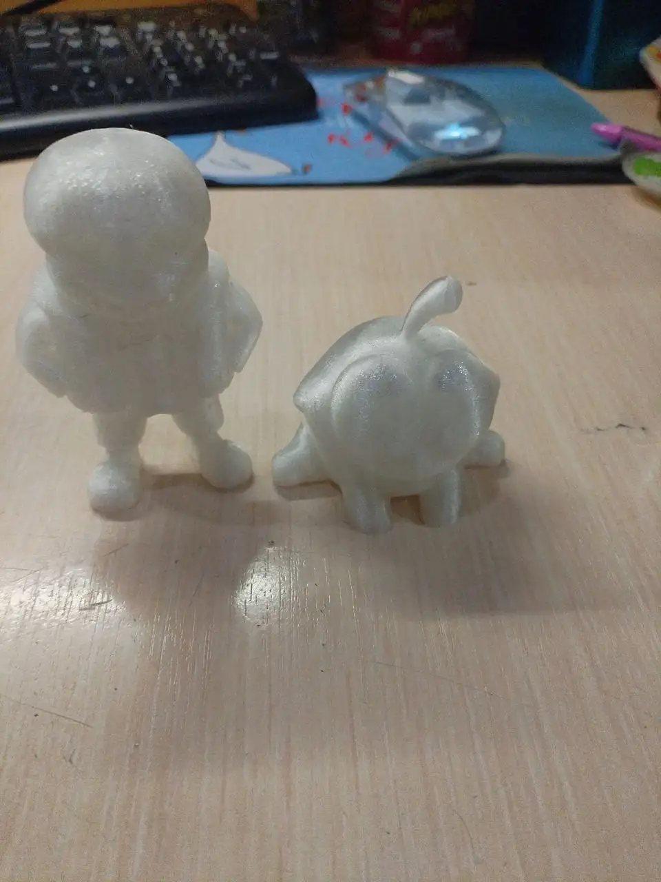 Two white 3D-printed figurines, one resembling a humanoid and the other a turtle-like creature, stand on a wooden desk. The…