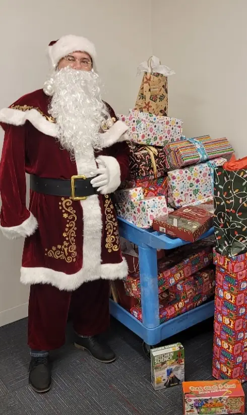 A man dressed as Santa Claus stands beside a blue cart piled high with colorful, wrapped Christmas presents. He wears a…