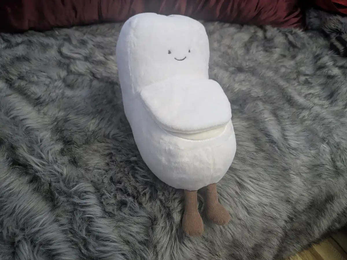 A white, plush, toilet-shaped stuffed toy with a smiling face and brown corduroy legs sits upright on a fluffy gray rug. The…