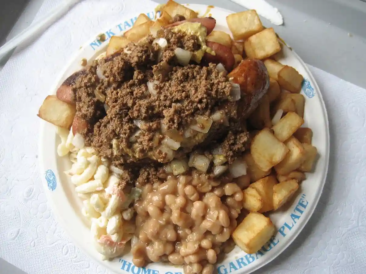 A plate piled high with a hot dog, baked beans, macaroni salad, and fried potatoes, all topped with a thick brown…