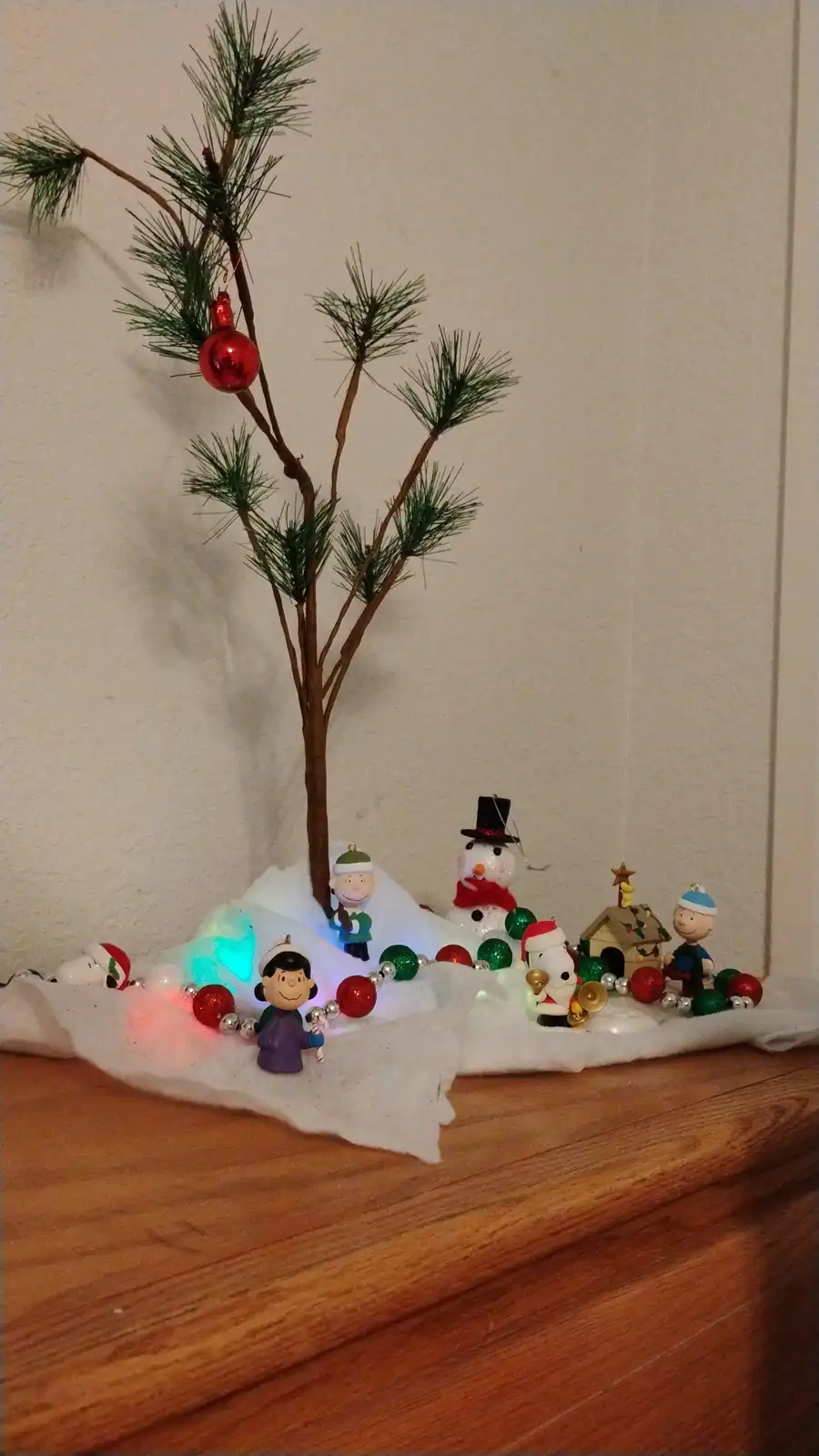 A small, sparse artificial pine tree with one red ornament sits on a bed of white faux snow on a wooden surface, surrounded…