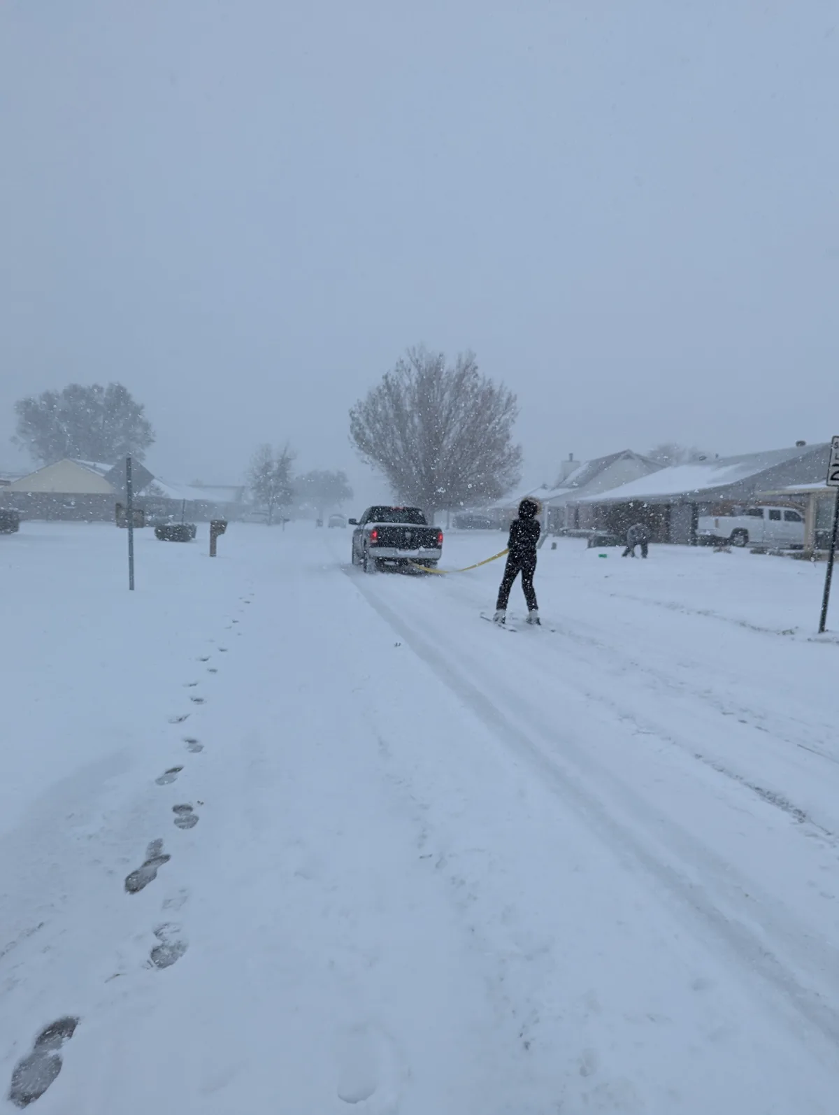 A person wearing dark winter clothing and skis is being pulled by a black pickup truck through a snow-covered residential…