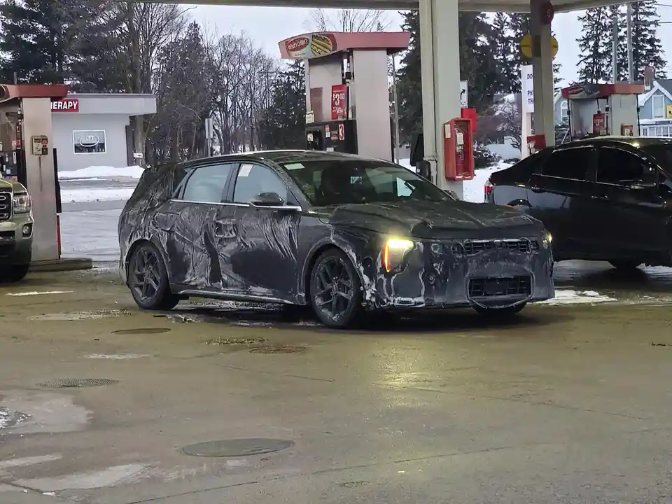 A dark-colored car, covered in black camouflage wrap typically used for spy photos, is parked at a gas station. The car's…