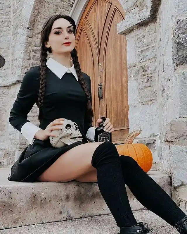 A woman dressed as Wednesday Addams sits on stone steps in front of a wooden door, holding a skull and a bottle labeled…