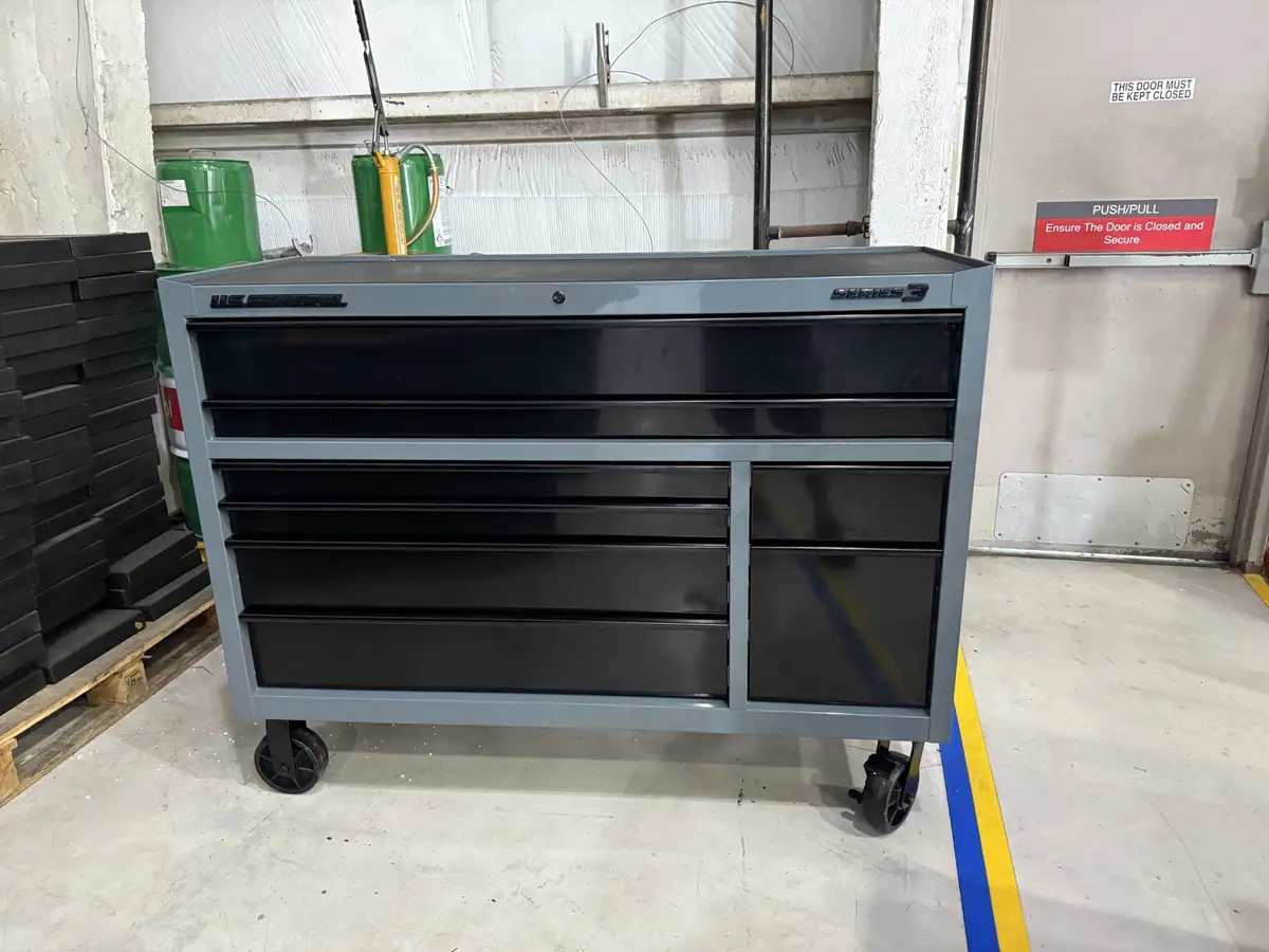 A large, grey, wheeled U.S. General Series 3 tool chest with multiple black drawers sits in a warehouse, surrounded by green…