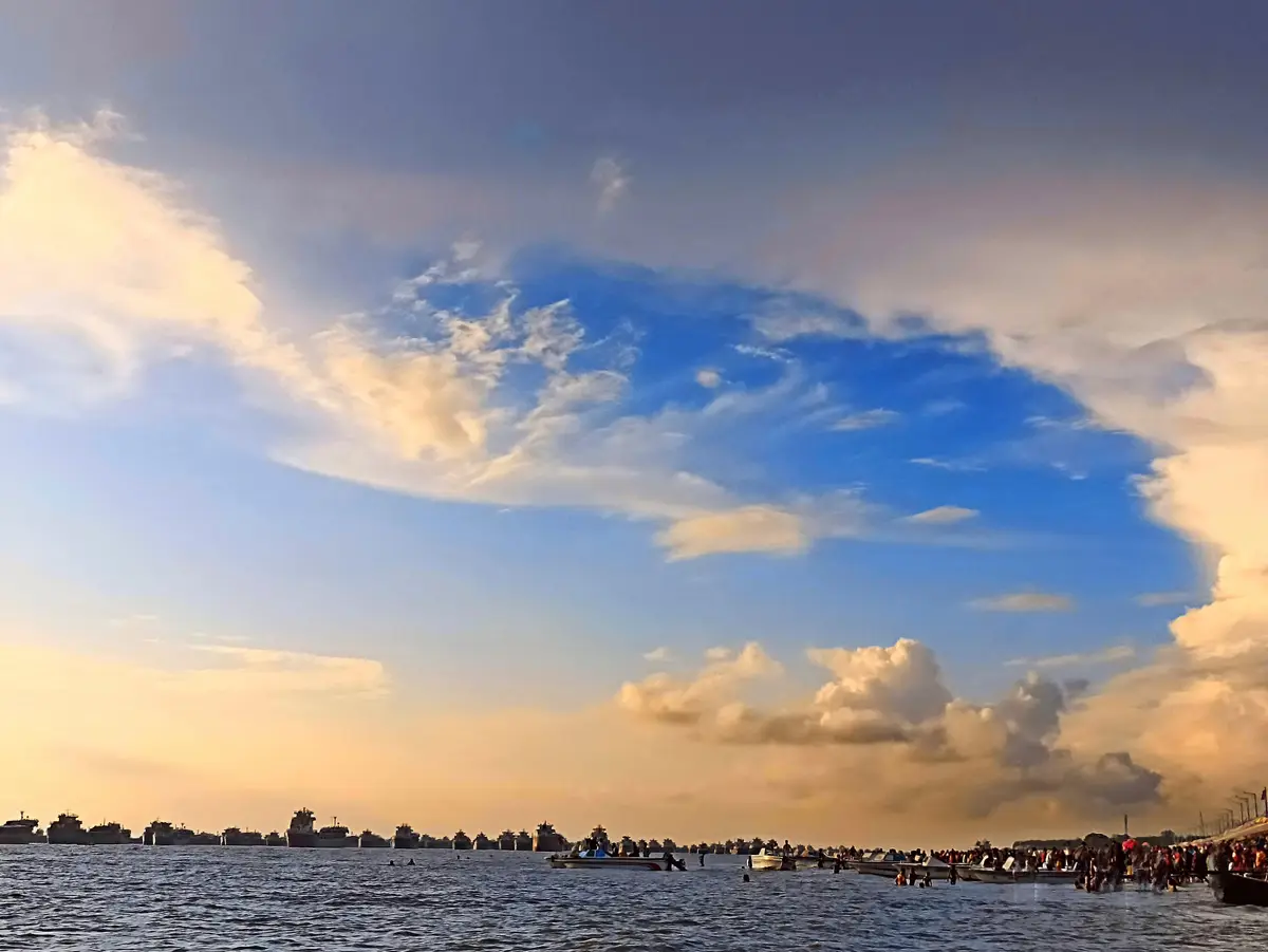 A scenic photo of a crowded waterfront at sunset with large ships in the distance and people in small boats and shallow…