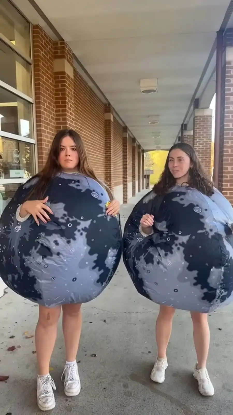 Two young women stand side-by-side on a covered school walkway, each wearing a large, inflatable costume resembling the moon…