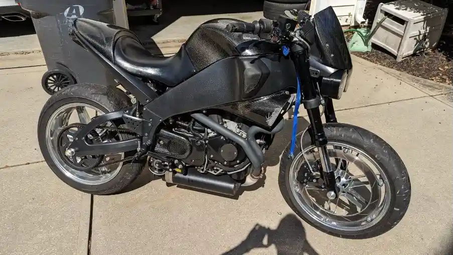 a black custom motorcycle with carbon fiber body panels and chrome wheels is parked on a concrete driveway. The bike has a…