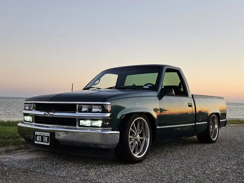 A dark green Chevrolet pickup truck with custom silver wheels is parked on gravel near a body of water during sunset. The…
