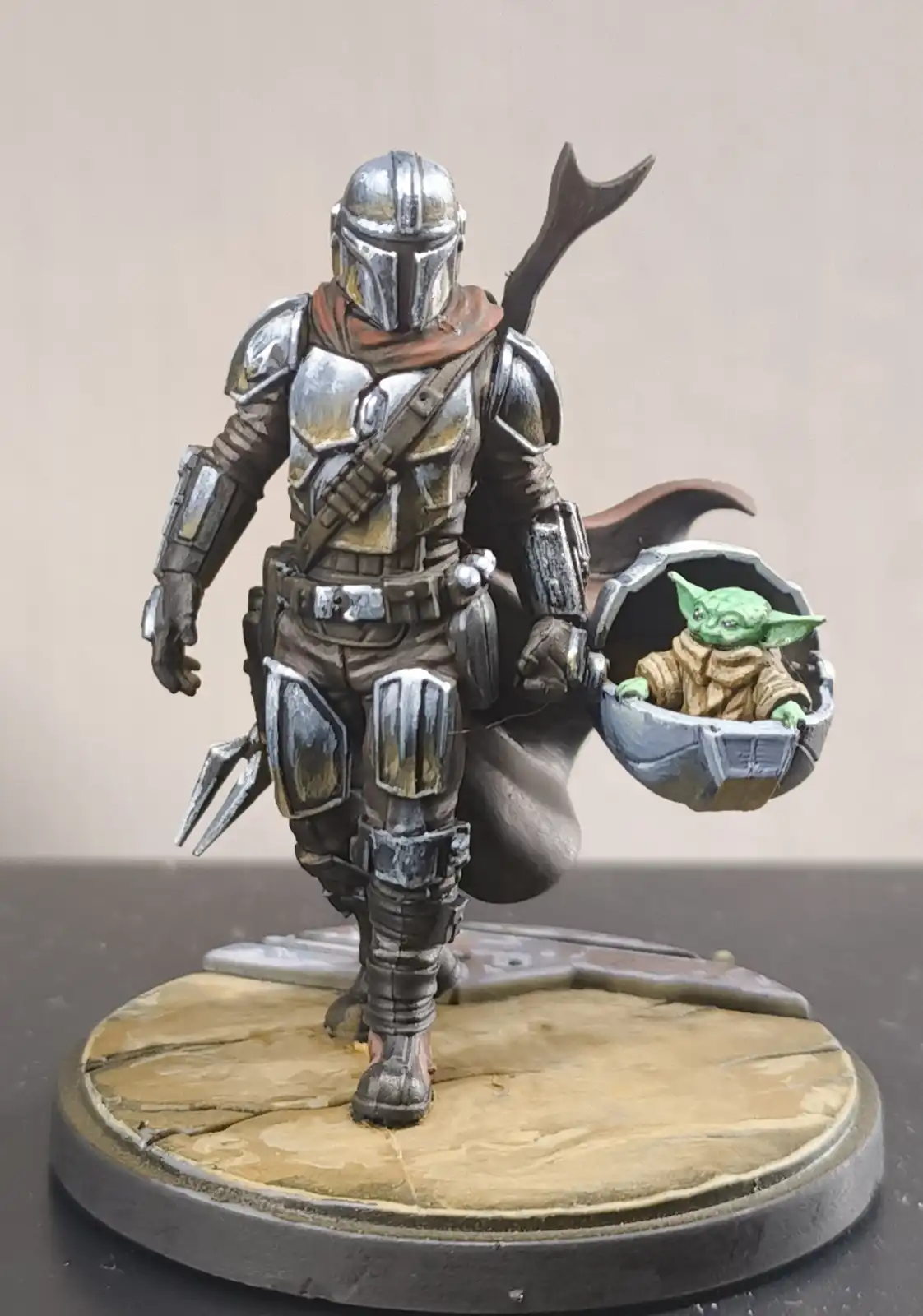 A painted miniature figure of The Mandalorian, wearing his iconic armor and carrying Grogu (Baby Yoda) in his hip-mounted…