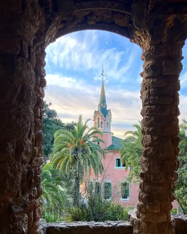A view through a rough stone archway frames a pink building with a colorful, ornate spire and green shutters, surrounded by…