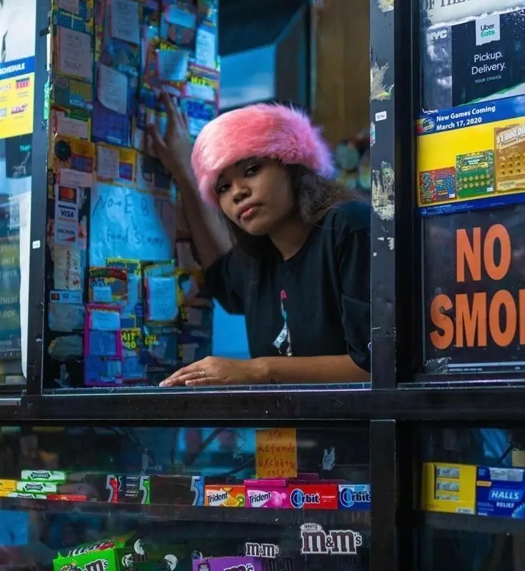 A woman wearing a bright pink fuzzy hat leans over a store counter, looking directly at the camera with a deadpan…