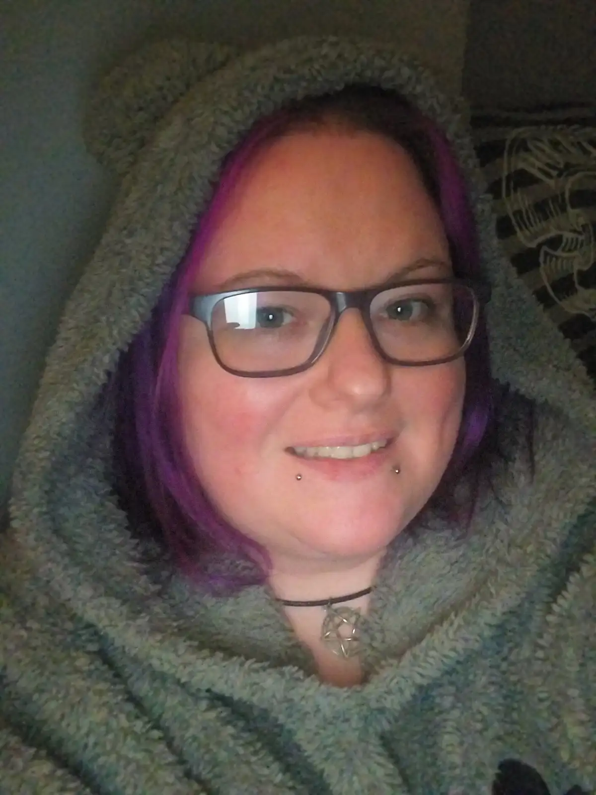 A person with purple hair and glasses smiles while wearing a fuzzy green hooded garment. They have a pentagram pendant…
