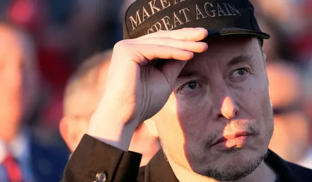 A close-up photo of Elon Musk wearing a black baseball cap with the text 'MAKE AMERICA GREAT AGAIN' embroidered in white. He…