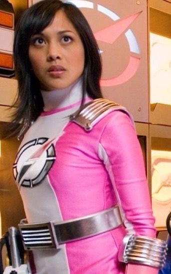 A woman in a pink and white superhero costume with silver accents and a belt, standing in a sci-fi setting, looks directly…
