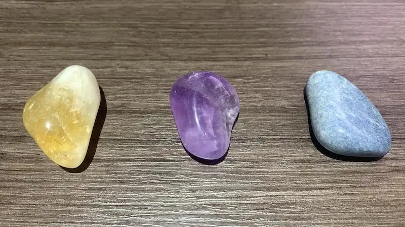 Three polished, smooth stones — one yellow, one purple, and one light blue — are arranged in a row on a wooden surface. The…