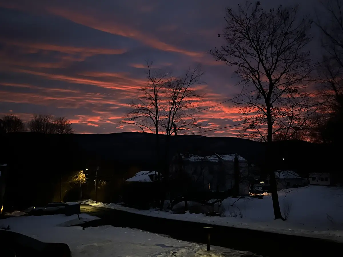 A snowy suburban street at dusk with silhouetted trees and houses against a dramatic sky painted with fiery orange and…