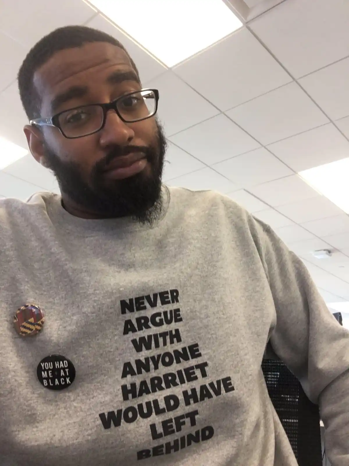 A man with glasses and a beard wears a gray sweatshirt with bold black text reading 'NEVER ARGUE WITH ANYONE HARRIET WOULD…